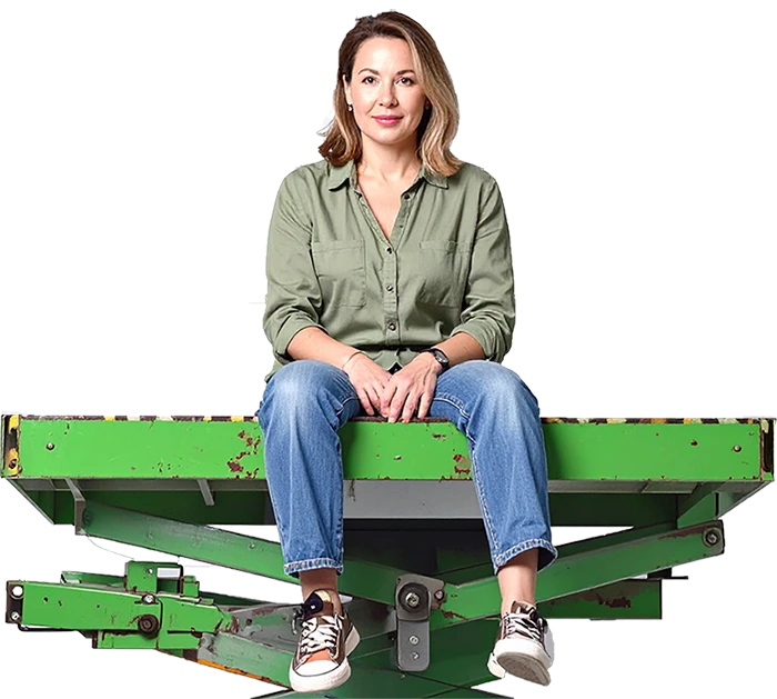 Frau sitzt auf grüner Plattform, trägt olivgrünes Hemd, Jeans und Sneakers, neutraler Hintergrund.