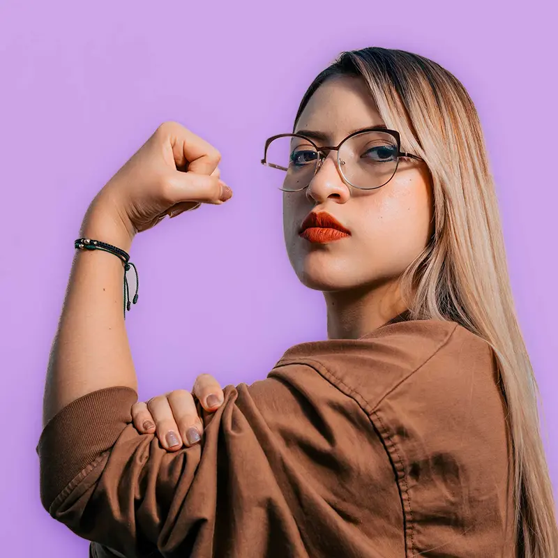 Selbstbewusste junge Frau mit Brille zeigt Stärke-Geste vor lila Hintergrund.