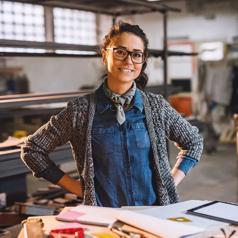 Frau mit Brille und Halstuch steht in Werkstatt und lächelt selbstbewusst in die Kamera.