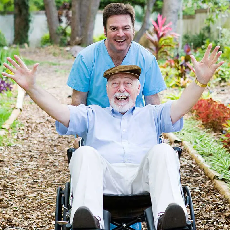 Pfleger schiebt lachenden Mann im Rollstuhl durch einen Gartenweg.