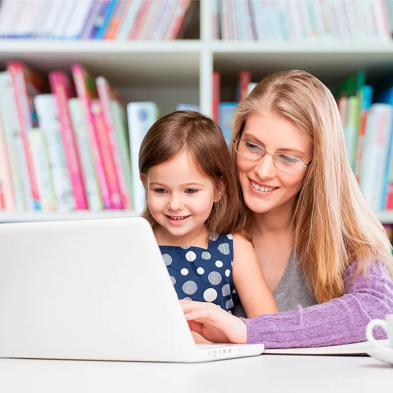 Lächelnde Mutter lernt mit ihrer Tochter am Laptop vor einem Bücherregal.