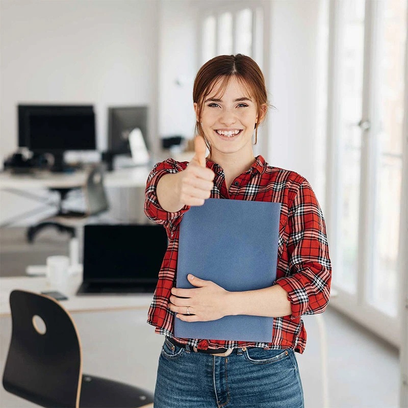 Junge Frau im Büro zeigt Daumen hoch und hält eine blaue Mappe, lächelnd in die Kamera.