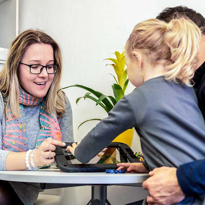 Frau zeigt Kind ein Tablet, während ein Erwachsener daneben sitzt.